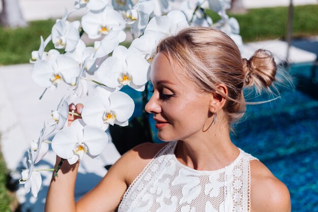 Outdoor portrait of woman in white wedding dress sitting near blue swimming pool with flowers