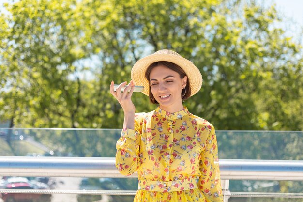 Outdoor portrait of woman in yellow summer dress and hat listen audio voice message on phone