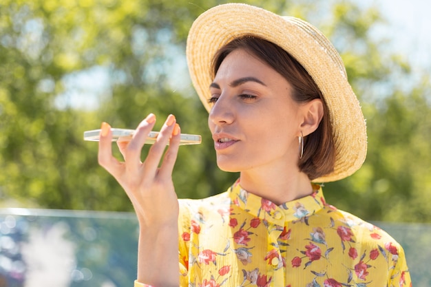 Outdoor portrait of woman in yellow summer dress and hat recording audio voice message on phone