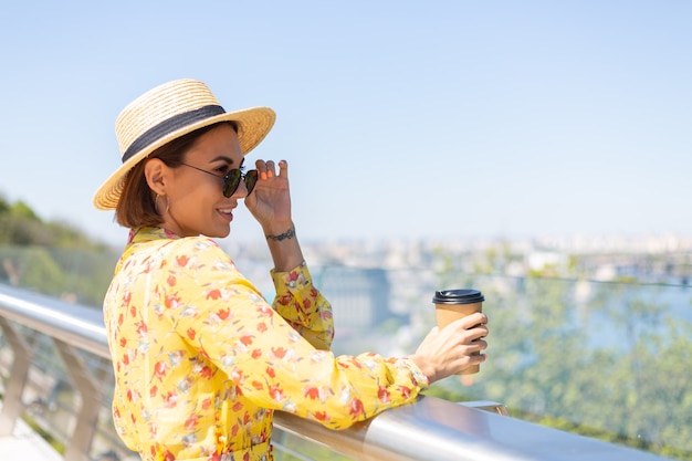 太陽を楽しんでいるコーヒーのカップと黄色の夏のドレスと帽子の女性の屋外の肖像画は、街の素晴らしい景色と橋の上に立っています