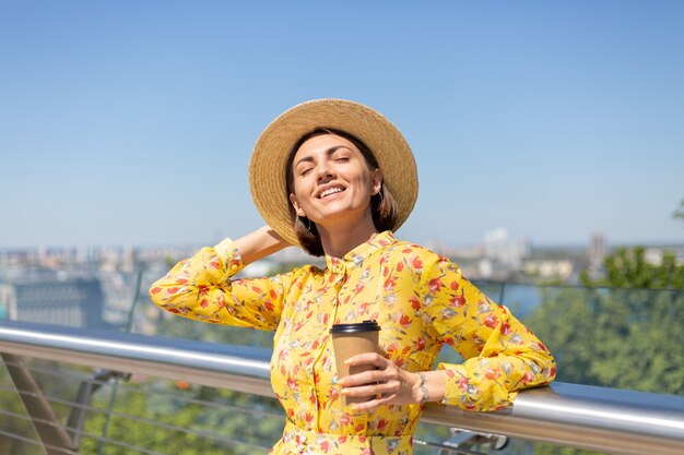 太陽を楽しんでいるコーヒーのカップと黄色の夏のドレスと帽子の女性の屋外の肖像画は、街の素晴らしい景色と橋の上に立っています