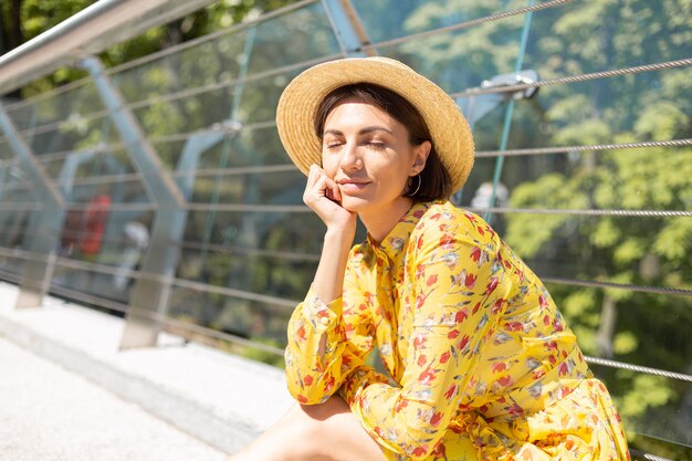 Outdoor portrait of woman in yellow summer dress sitting on bridge with eyes closed, happy mood, enjoying sunny summer days