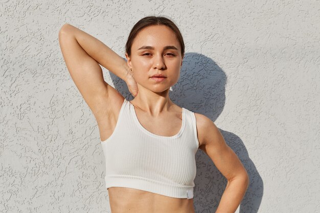 Outdoor portrait of a young athletic girl posing near white wall with raised arm, looking at camera, being photographed during workout, healthy lifestyle.