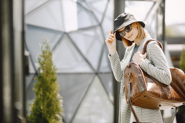 大きな茶色の革のバッグを持って、通りを歩いている若い美しいファッショナブルな女性の屋外の肖像画。サングラスと黒い帽子をかぶったブロンドの女の子