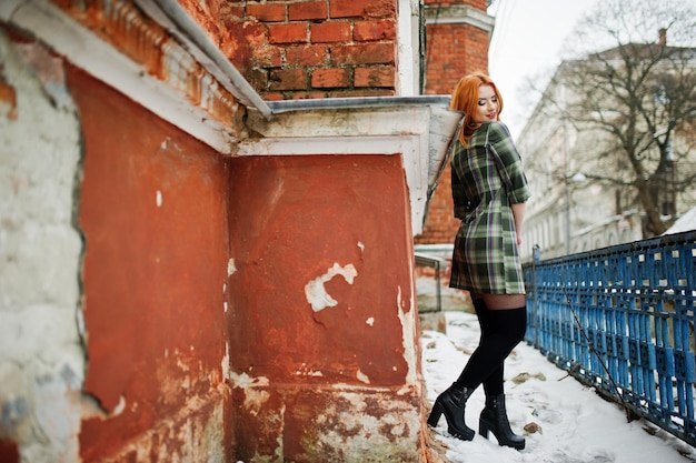 An outdoor portrait of a young pretty girl with red hair wearing checkered dress with girly backpacks standing on the brick wall background in winter day