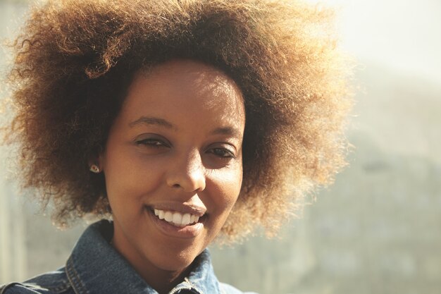 Outdoor portrait of young stylish dark-skinned female with curly hair wearing denim jacket, smiling, posing and enjoying sunny weather during morning walk.