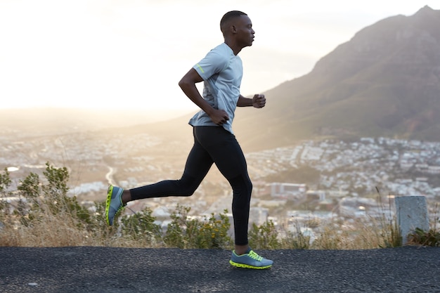 Outdoor shot of active dark skinned man running at morning, has regular trainings, dressed in tracksuit and comfortable sneakers, concentrated into distance, sees finish far away.