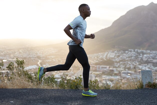 Outdoor shot of active dark skinned man running at morning, has regular trainings, dressed in tracksuit and comfortable sneakers, concentrated into distance, sees finish far away.