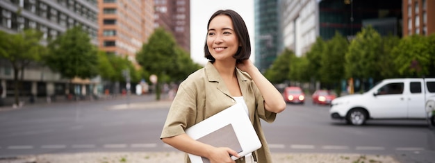 Free photo outdoor shot of asian girl with laptop going somewhere in city centre walking on street going to