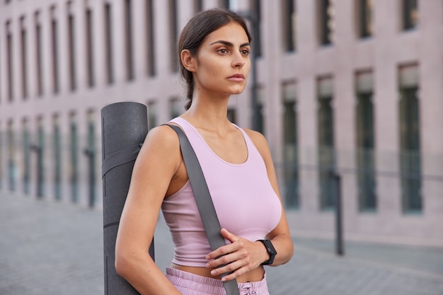 Outdoor shot of attentive young fitness model prepares for pilates exercising carries karemat looks forwards thinks about healthy lifestyle strolls in modern city admires views feels motivated