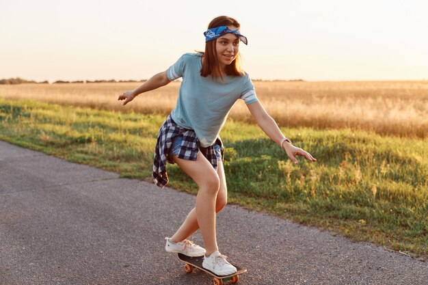 Outdoor shot of attractive female wearing casual style clothing riding skateboard on street, looking away with smile and concentrated expressing, being happy of her achievement.