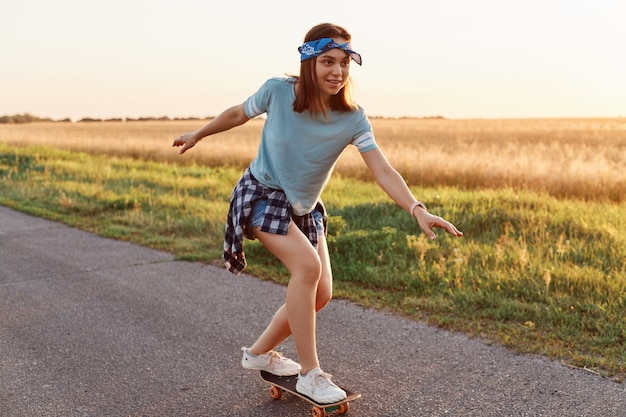 캐주얼 스타일의 옷을 입고 거리에서 스케이트보드를 타고 있는 매력적인 여성의 야외 사진, 미소로 시선을 돌리고 집중적인 표현, 그녀의 성취에 만족합니다.