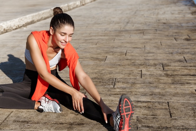 Снимок привлекательной фитнес-женщины, которая выглядит счастливой и растягивает ногу перед тренировкой, бегущей трусцой ...