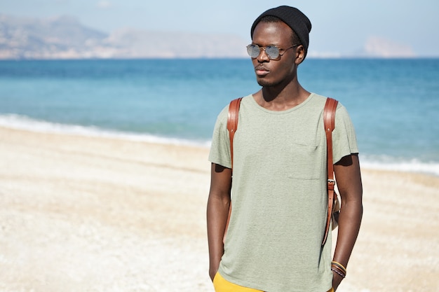 Outdoor shot of attractive serious African male wearing round shades and hat spending vacations by the sea