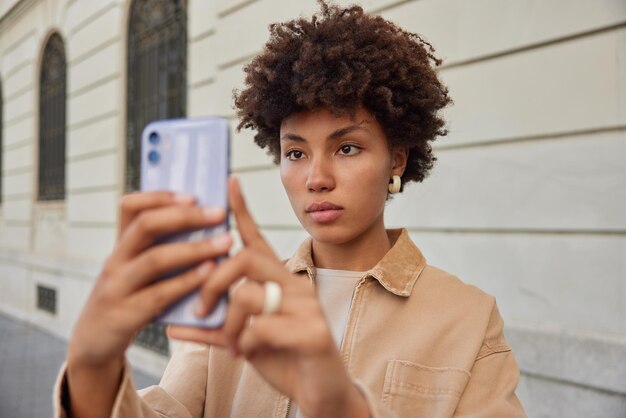 Outdoor shot of attractive woman with curly hair uses front mobile camera for clicking selfie images wears stylish clothes shoots influence video vlog during leisure time stands near city building
