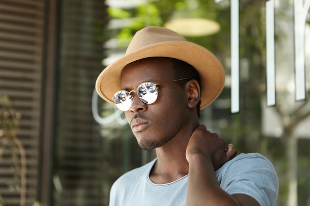 Outdoor shot of attractive young African American male tourist hiding from scorching rays of summer sun in shadow of urban cafe, dressed in stylish clothing, looking frustrated, rubbing his neck