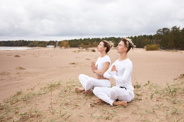 Free photo outdoor shot of attractive young woman and man dressed in similar white clothes sitting barefooted on deserted sandy beach with legs crossed, meditating, keeping eyes closed, making namaste gesture