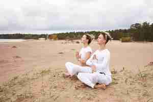 Free photo outdoor shot of attractive young woman and man dressed in similar white clothes sitting barefooted on deserted sandy beach with legs crossed, meditating, keeping eyes closed, making namaste gesture