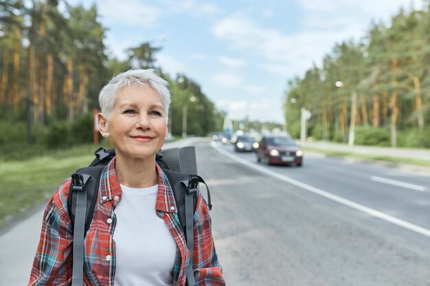 Outdoor shot of beautiful active middle aged woman with short haircut carrying backpack walking along high road while hitchhiking alone.