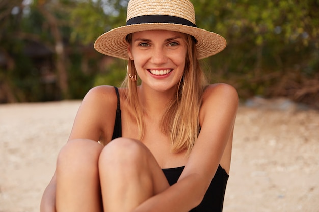 Outdoor shot of beautiful female wears straw hat and swimsuit, sits on hot sandy beach, being satisfied with weather conditions and good rest, feels happy and relaxed. Woman on sandy beach alone