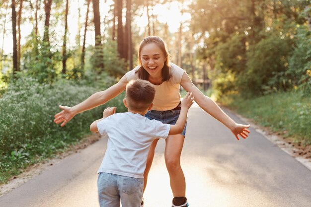 이빨 미소와 긍정적 인 행복식이있는 아름다운 여성의 야외 촬영은 여름 공원에서 함께 롤러 블레이드를 타는 동안 팔을 벌려 그녀의 아들을 잡는 긍정적 인 행복 식입니다.