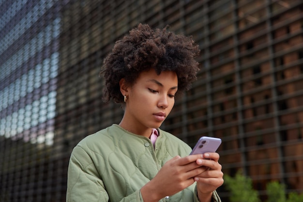 Foto gratuita scatto all'aperto di una bella donna concentrata sullo schermo dello smartphone che legge notizie o scrive messaggi di testo indossando la giacca