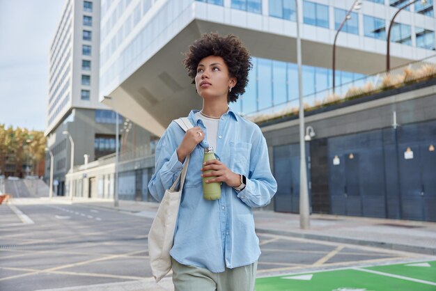 巻き毛の美しい若い女性の屋外ショットは頭上に見えますカジュアルなストリートの服を着てバッグとボトルを運びます。都会の環境で淡水散歩は晴れた日を楽しんでいますライフスタイルのコンセプト