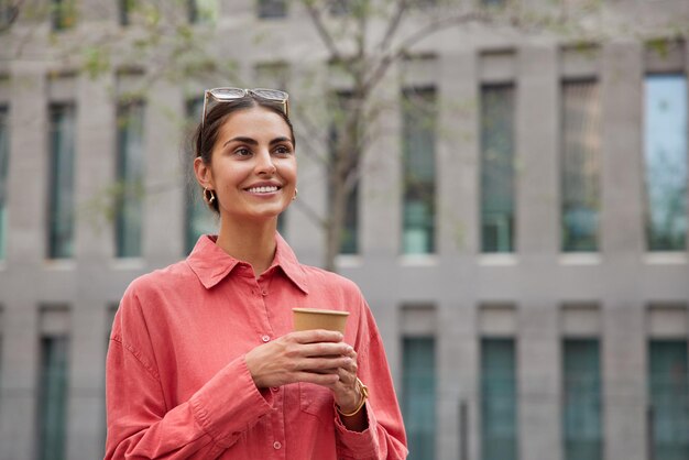 Outdoor shot of cheerful young European woman holds coffee to go dressed casually strolls against city building.