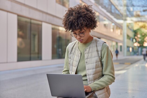 Outdoor shot of curly female student with curly hair dressed in casual clothes uses modern laptop prepares course work or new project poses in city Female entrepreneur works remotely outside