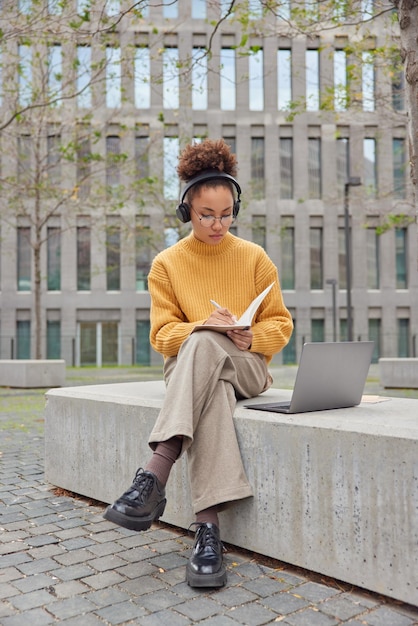 Outdoor shot of curly haired female student sits outdoors in front of opened laptop computer writes down notes in notepad prepares for lesson poses against modern city building E learning concept