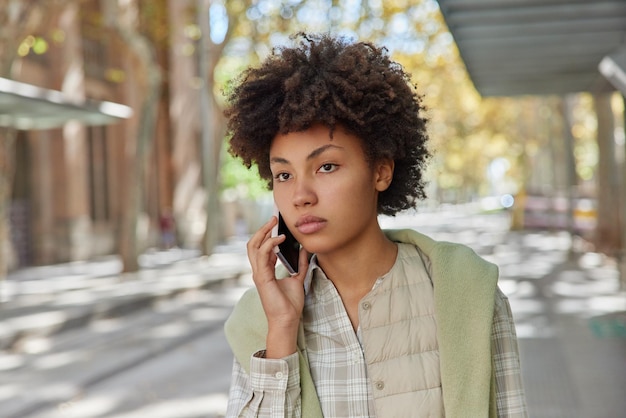 Outdoor shot of curly haired woman makes smartphone call phones friend looks thoughtfully forward dressed in casual clothes poses at street against blurred background Communication concept