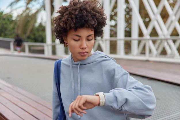 Outdoor shot of curly haired woman wears casual hoodie checks time on smartwatch carries bag waits for someone has serious expression waits for trainer to start sport training People and lifestyle