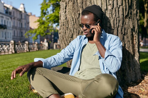 Outdoor shot of dark-skinned male in trendy glasses, hat, shirt and trousers, sitting crossed legs on green lawn near tree, spending his free time at park, speaking over smart phone with friend