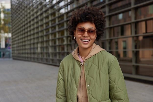 Outdoor shot of fashionable attractive woman smiles broadly wears sunglasses sweatshirt and jacket strolls in street of modern city being in good mood enjoys leisure time