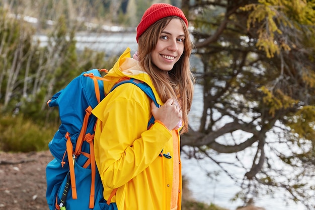 Outdoor shot of glad female with broad smile, stands sideways at camera, carries rucksack