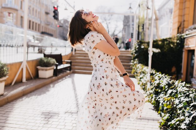 Outdoor shot of graceful girl relaxing in sunny day. Stylish short-haired lady in white dress dancing on the street.