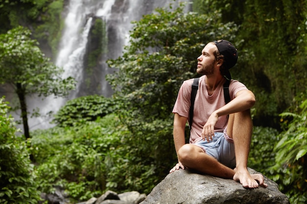 Outdoor shot of handsome barefooted young traveler with beard having rest on big rock during his hiking trip in rainforest
