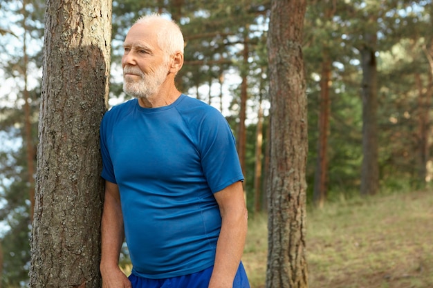 Colpo all'aperto dell'uomo caucasico barbuto anziano bello che indossa la maglietta asciutta blu adatta che posa in legno, spalla pendente sul pino, avendo riposo dopo l'allenamento cardio mattutino, ammirando il bellissimo paesaggio