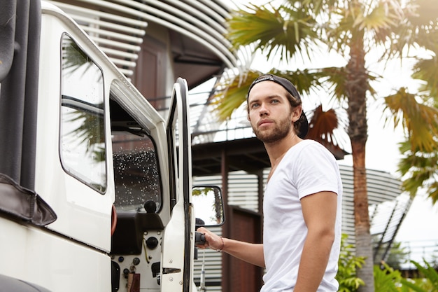 Free photo outdoor shot of handsome young bearded man in t-shirt standing outside his white crossover utility vehicle, pulling its door handle and looking away, ready to get in