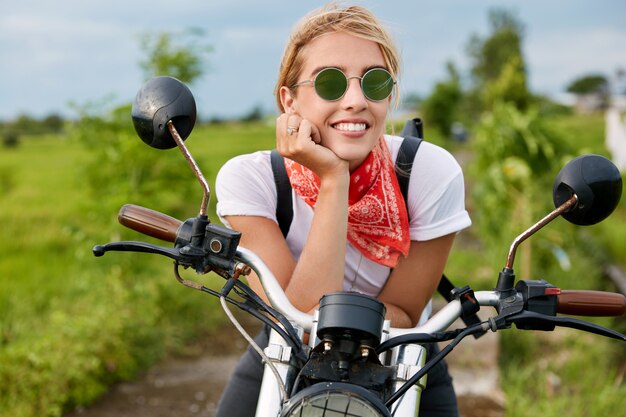 Outdoor shot of happy blonde female motorcyclist wears casual t shirt and sunglasses, look into distance with cheerful expression, sits on motorbike, poses in countryside. Traveling and freedom