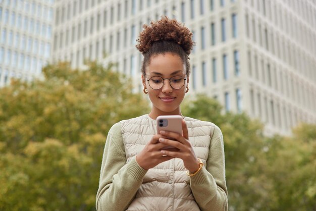 幸せな巻き毛の若い女性の屋外ショットは、スマートフォンでニュースを読むオンライン市場で割引を楽しんでいます