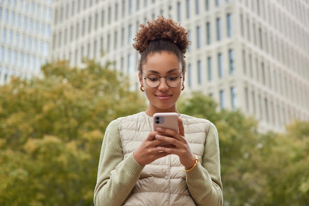 Outdoor shot of happy curly haired young woman enjoys discounts in online market reads news on smartphone