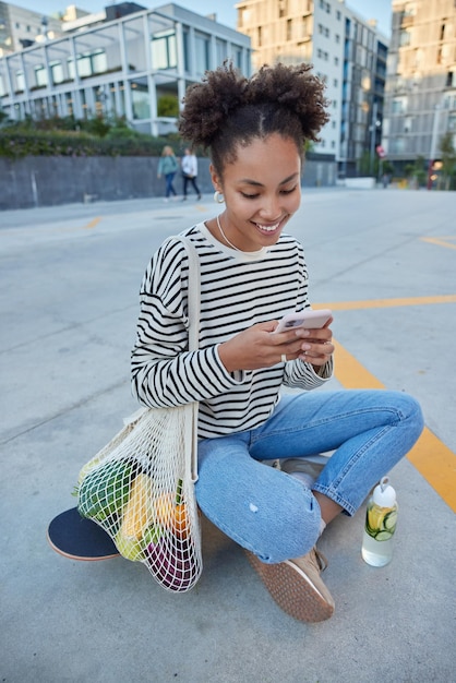 幸せな縮れ毛の若い女性の屋外ショットは、携帯電話を使用してテキストメッセージを送信しますストライプのジャンパーを着用し、ジーンズはスケートボードに座って市内中心部のロングボードのコンセプトで嬉しい表情のポーズをとっています