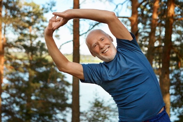Outdoor shot of happy energetic senior retired man enjoying physical training in park, doing side bends exercise, holding hands together with broad smile, warming up body before run