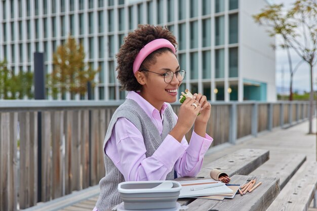 Outdoor shot of happy femaleeats delicious sandwich draws in notebook with crayons works on creative project creats pictures wears transparent glasses elegant shirt and vest poses against urban area