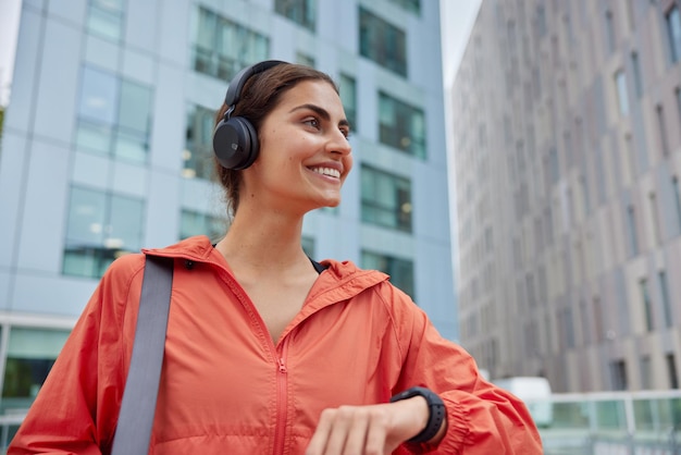 Outdoor shot of happy satisfied woman in anorak glad to monitor fitness results on smartwatch listens music from playlist has glad expression poses against blurred background. Sporty lifestyle