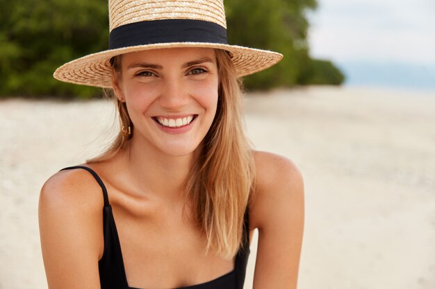 Outdoor shot of happy young woman with long hair, sunbathes on tropical beach, wears straw hat, being satisifed after swimming or walking on coastline. Leisure in summer and happiness concept