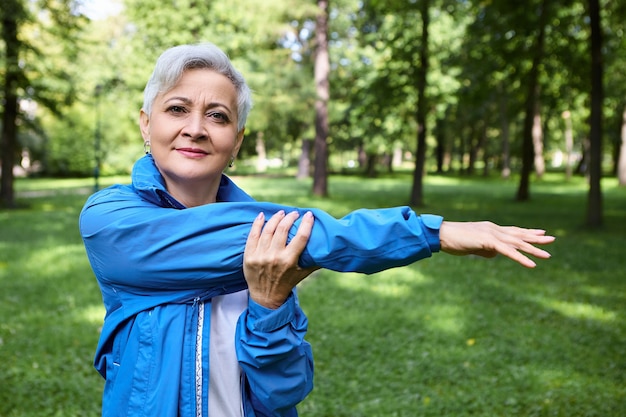 Outdoor shot of healthy sporty elderly woman with short gray hair exercising in park. Senior female in blue sports jacket stretching arm muscle, warming up before running workout