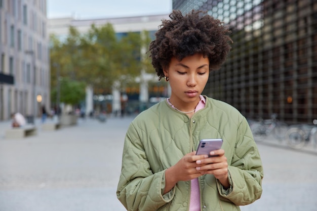 무료 사진 곱슬머리를 한 진지한 여성의 야외 사진은 휴대폰 화면에 초점을 맞춘 스마트폰 유형 sms를 사용하여 낮 동안 거리에서 재킷을 입고 산책합니다. people 현대 기술 및 라이프스타일 개념