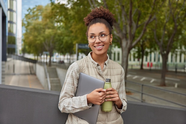 Outdoor shot of pensive cheerful young woman looks around while walking in city carries digital tablet and bottle of fresh water